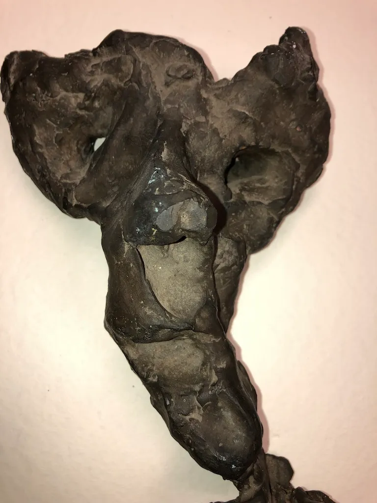 A dark, roughly modelled torso mounted on a pale wall, its surface broken into hollows and ridges and tapering to a single leg.