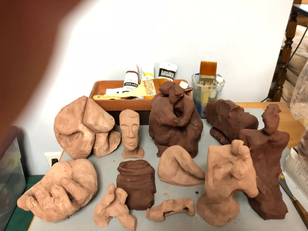 The studio table seen wider, a dozen clay pieces gathered on grey paper.