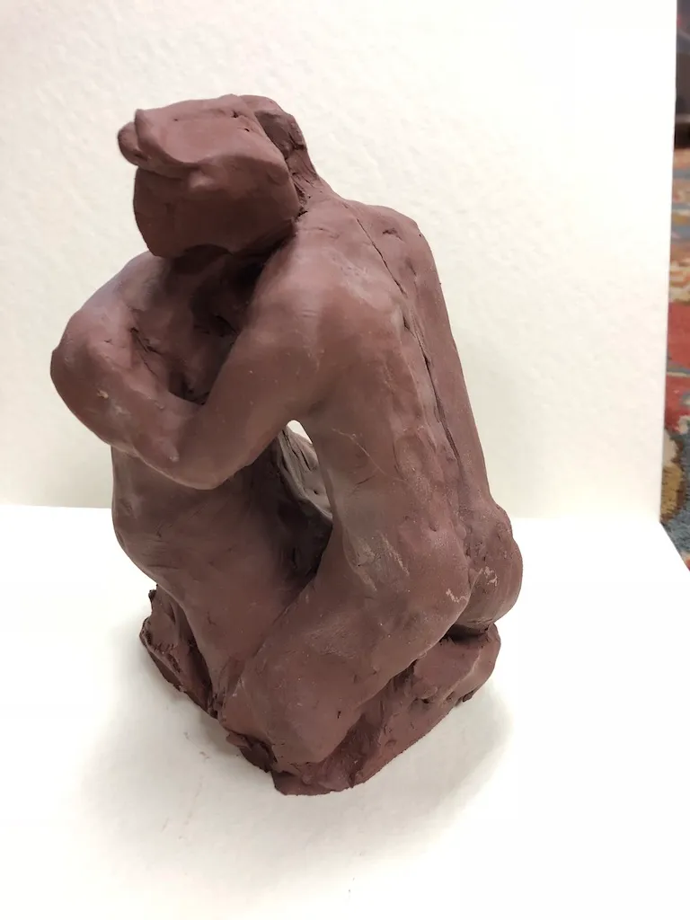 A red-brown clay figure kneeling and bent forward, arms wrapped around itself and its head lowered.