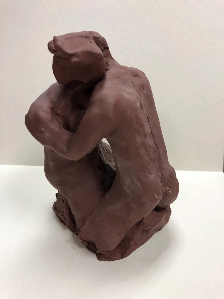 A red-brown clay figure kneeling, arms folded over its drawn-up knees.