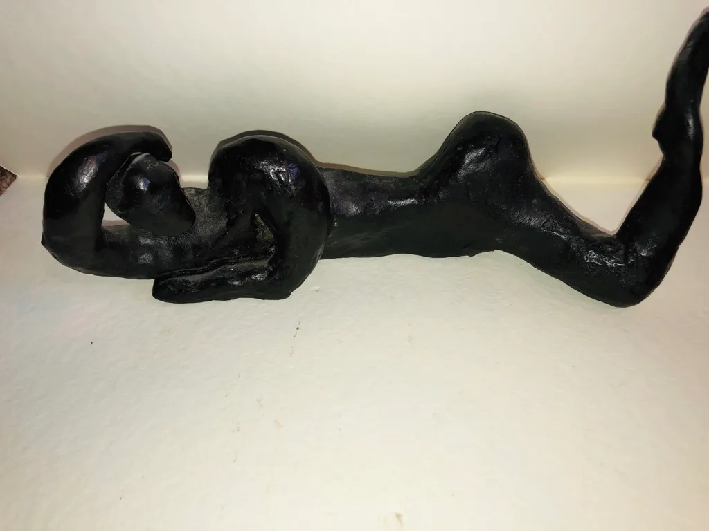 A black clay figure lying on its back, one arm curled beneath its head and one leg lifted.